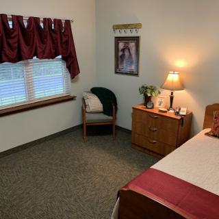 A cozy bedroom featuring a bed, a wooden dresser, a lamp, and a chair by the window.