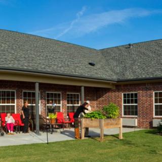 A group of people gather in the backyard of a brick house with a raised garden bed and outdoor seating.