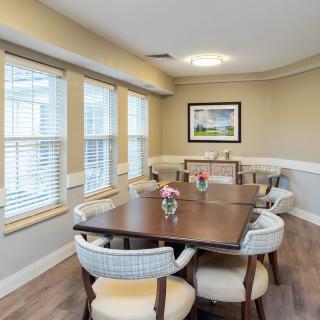 A cozy dining room with a large wooden table and upholstered chairs, accented by flowers and natural light streaming through multiple windows.