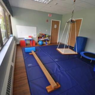 A therapy room equipped with gym equipment and children's toys, featuring a swing, balance beam, trampoline, and blue floor mats.