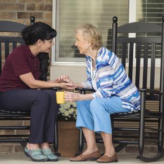 A caregiver and an elderly woman share a warm conversation on rocking chairs outside.