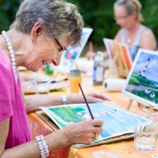 An elderly woman smiling as she paints during an outdoor art class.