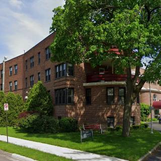 Brick apartment building in a residential neighborhood with trees and a sidewalk.