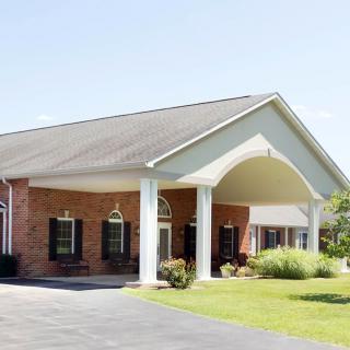 A building with a brick exterior, featuring a large covered entrance supported by columns and surrounded by a paved driveway and landscaping.