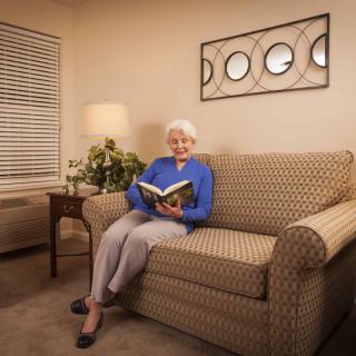 An elderly woman sitting on a sofa in the living room, reading a book with a warm lamp lighting the space.