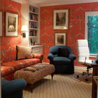 A cozy living room with red floral wallpaper, featuring a sofa, armchair, and home office corner.