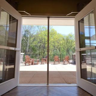 A view through open double doors revealing a sunny patio with outdoor seating and trees in the background.