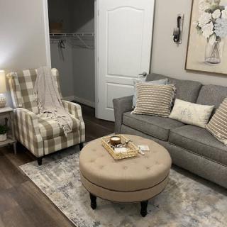 A cozy living room with a gray sofa, checkered armchair, and a beige ottoman atop a decorative rug.