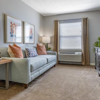 A cozy living room with a light blue sofa, decorative pillows, and framed artwork on the wall. The room is well-lit with a table lamp beside the sofa and natural light from a large window with curtains.