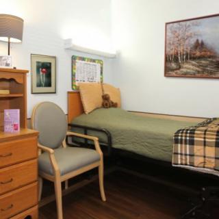 A cozy bedroom with a single bed, chair, and dresser, featuring warm lighting and a scenic painting on the wall.