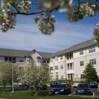 A residential apartment complex surrounded by blooming trees and a parking lot with cars under a cloudy sky.