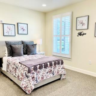 A cozy bedroom with a neatly made bed, adorned with decorative pillows and a floral blanket. The walls feature framed artwork, and a window allows natural light to fill the space.