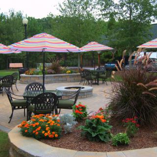 A well-landscaped outdoor patio area with tables, chairs, and umbrellas, surrounded by vibrant flowers and featuring a central fire pit.
