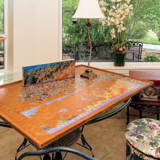A jigsaw puzzle in progress on a wooden table near a lamp and window, surrounded by indoor plants.