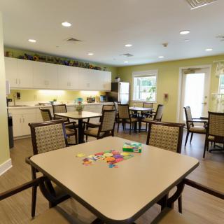 A well-lit dining and activity room with multiple tables and chairs, featuring a kitchenette area.