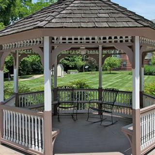 A wooden gazebo in a lush garden setting, featuring chairs inside and a well-manicured lawn in the background.