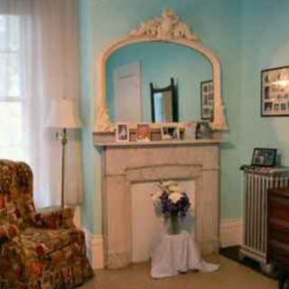 A cozy living room features a classic fireplace with an ornate mirror above it, blue walls, and a floral patterned chair.