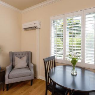 A cozy living room corner featuring a comfortable chair, a small round table, and a window with shutters. An air conditioner is mounted on the wall, and a vase of flowers adorns the table.