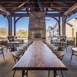 A modern outdoor dining area with a long wooden table, surrounded by chairs, and featuring a central fireplace.