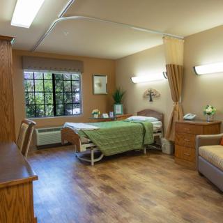 A well-lit hospital room featuring a single bed, wooden furniture, and a large window providing natural light.