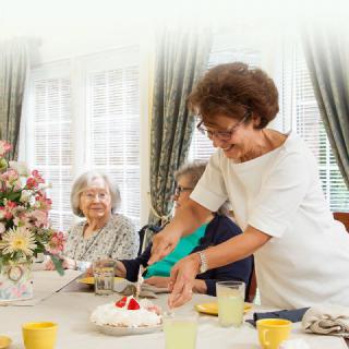 A group of elderly women enjoying a gathering, with one woman cutting a cake and smiling.