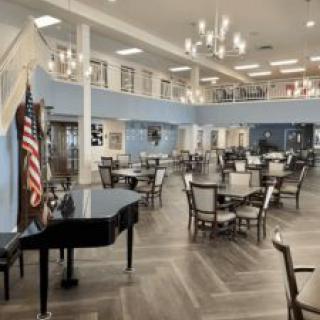 A spacious dining hall with elegant chandeliers and a grand piano, featuring neatly arranged tables and chairs.