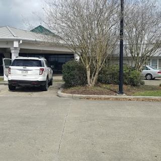 A view of a parking lot with two cars parked outside a building, surrounded by trees under a cloudy sky.