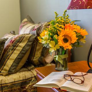A cozy bedroom corner featuring a vibrant floral arrangement, a book, and reading glasses.