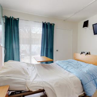 A well-lit hospital room featuring a bed with blue bedding, curtains, and a television.