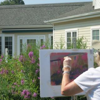 An artist painting outdoors in a garden setting, with a house in the background.
