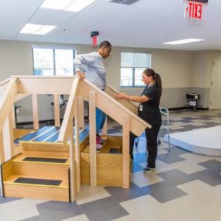 A person receives assistance during physical therapy, practicing walking on stairs indoors.