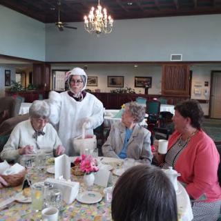 A group of people gathered around a dining table indoors, with a chandelier overhead.