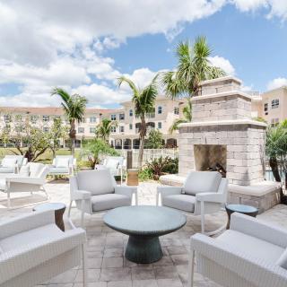 A stylish outdoor patio with modern furniture and a stone fireplace, surrounded by palm trees and buildings under a cloudy sky.