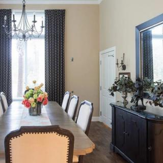 A stylish dining room featuring a long table with chairs, a chandelier, a vase of flowers, and large curtains.
