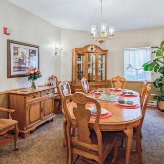 A cozy dining room featuring a wooden table set with red placemats, surrounded by matching chairs. A china cabinet stands against the wall, and a potted plant adds a touch of greenery to the space.