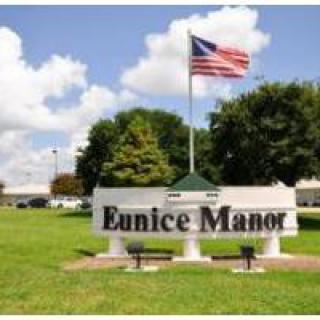 A sign for Eunice Manor stands in front of a grassy area with trees and an American flag.