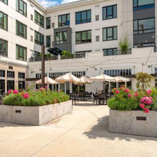 A modern outdoor patio with seating and umbrellas, surrounded by a multi-story building and flower planters.