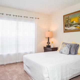A cozy bedroom with a neatly made bed, decorative pillows, a side table with a lamp, and a landscape painting on the wall.