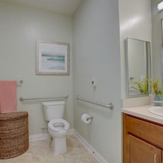 A clean and modern bathroom with light green walls, featuring a toilet, sink, mirror, towel basket, and wall artwork.