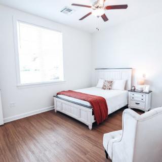 A bright and minimalist bedroom with wooden flooring, featuring a neatly made bed adorned with a red blanket and white furniture.