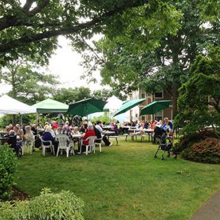 A group of people enjoying an outdoor event in a garden setting with tables, chairs, and umbrellas.