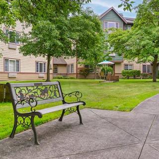 A peaceful outdoor area with a bench and pathway near an apartment building, surrounded by trees and grass.