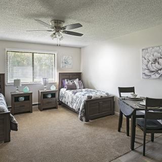 A spacious bedroom with two twin beds, each accompanied by a nightstand. The room features a large window for natural light, a ceiling fan, and a small dining table with chairs.
