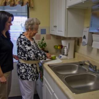 A caregiver assists an elderly woman in the kitchen as part of daily living activities.