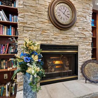 A cozy living room with a stone fireplace, large clock, bookshelf filled with books, and a vase of colorful flowers.