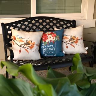 A stylish outdoor bench adorned with decorative floral and bird-themed cushions, perfect for a cozy patio setting.