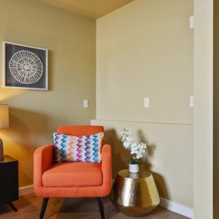 A cozy living room corner featuring an orange armchair with a patterned pillow, a decorative side table, and wall art.
