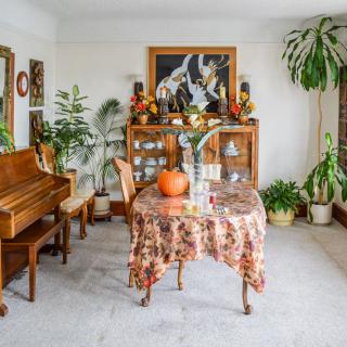 A cozy living room with a piano, house plants, and a table decorated with a pumpkin.