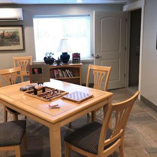 A cozy room featuring a table set with a chessboard and surrounded by chairs, a bookshelf beneath a window, and artwork on the wall.