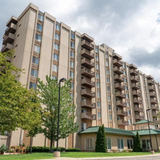 A tall residential apartment building with balconies, surrounded by trees under a cloudy sky.